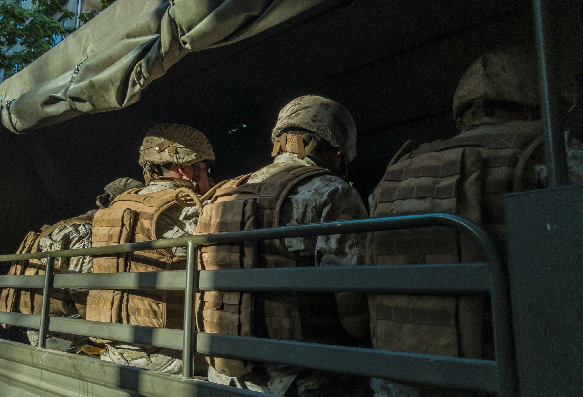 Service members in helmets and tactical vests, seen from behind, seated in the back of a military truck.