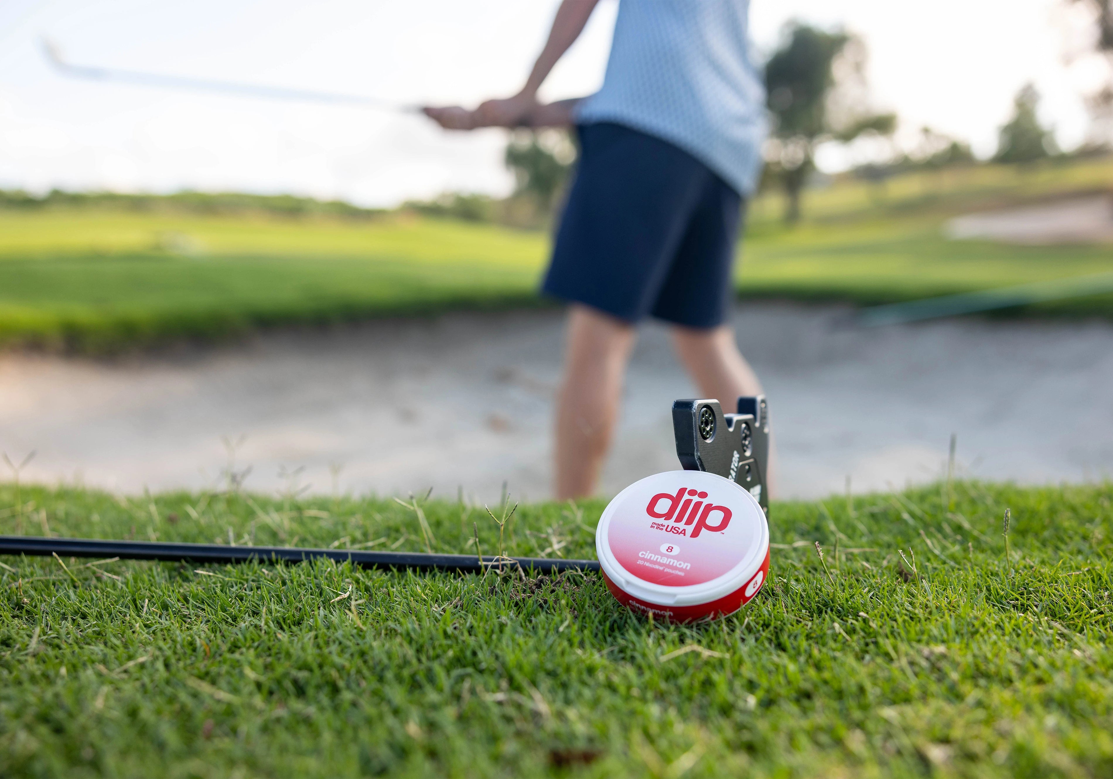 'diip' branded 8 cinnamon nicotine-free dip pouch can laying next to a golf club and a man golfing on a golf course in the background 