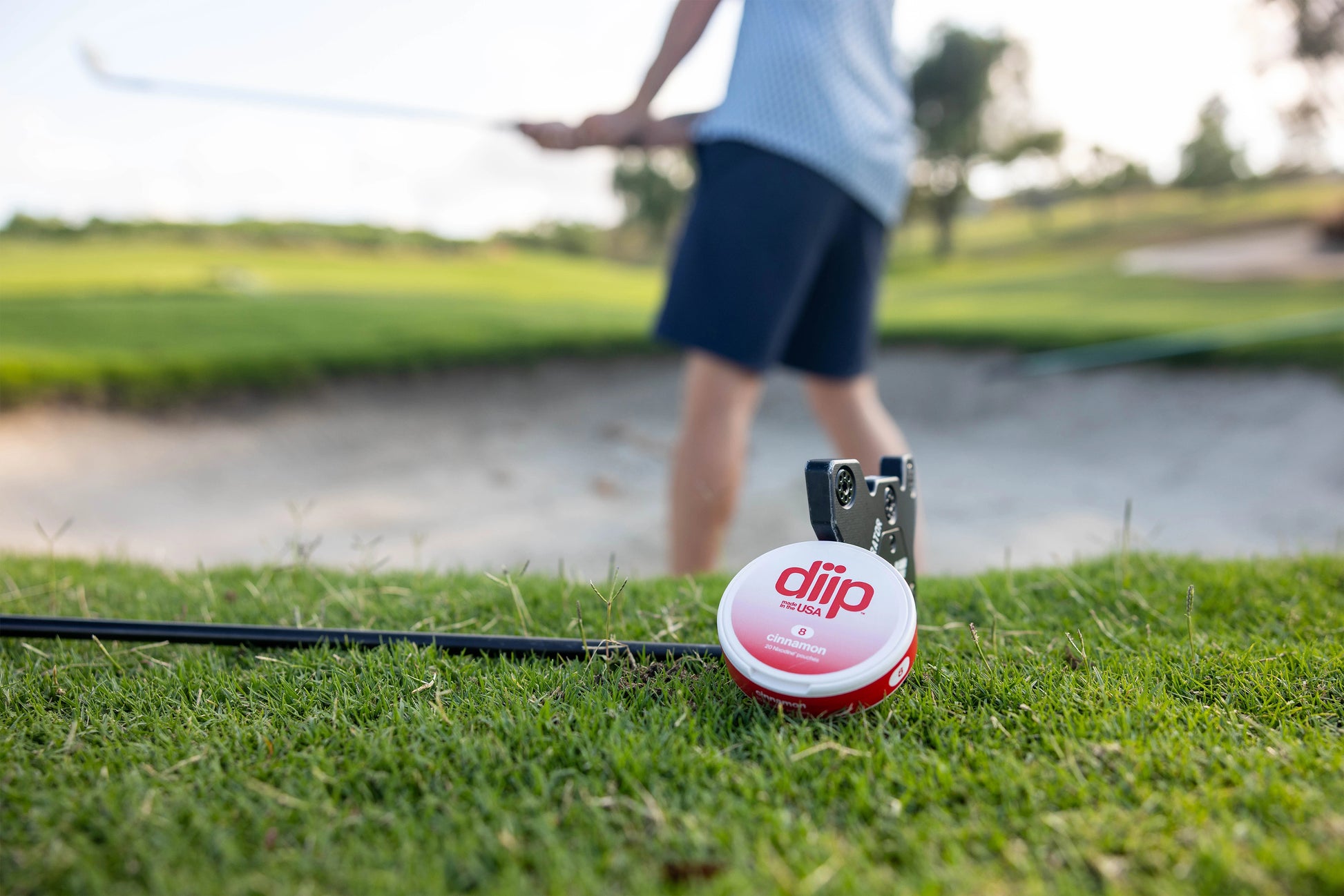 'diip' branded 8 cinnamon nicotine-free dip pouch can laying next to a golf club and a man golfing on a golf course in the background 