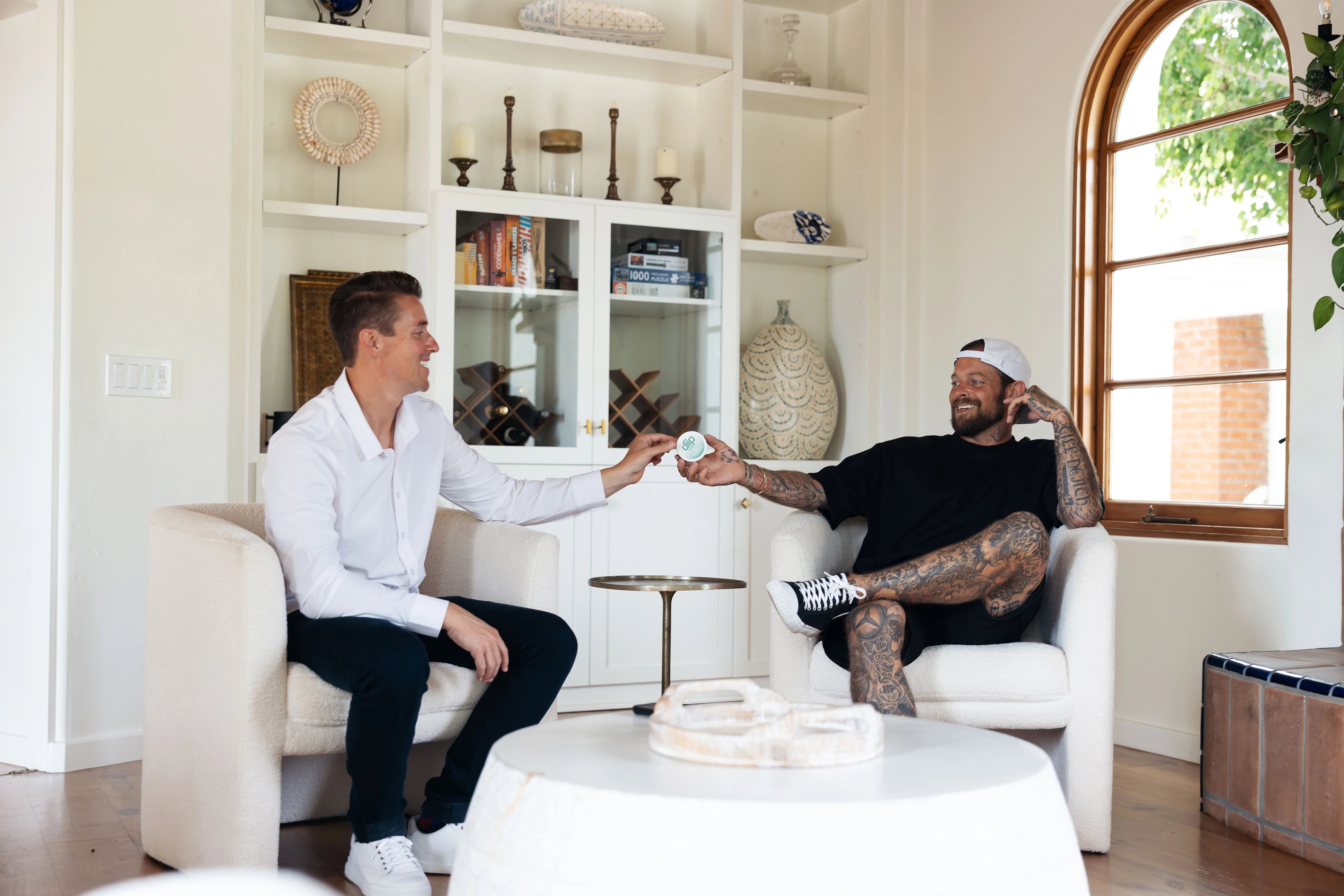 Two men sitting in a modern living room with a white bookshelf and large window holding a 'diip' branded wintergreen nicotine-free dip pouch can