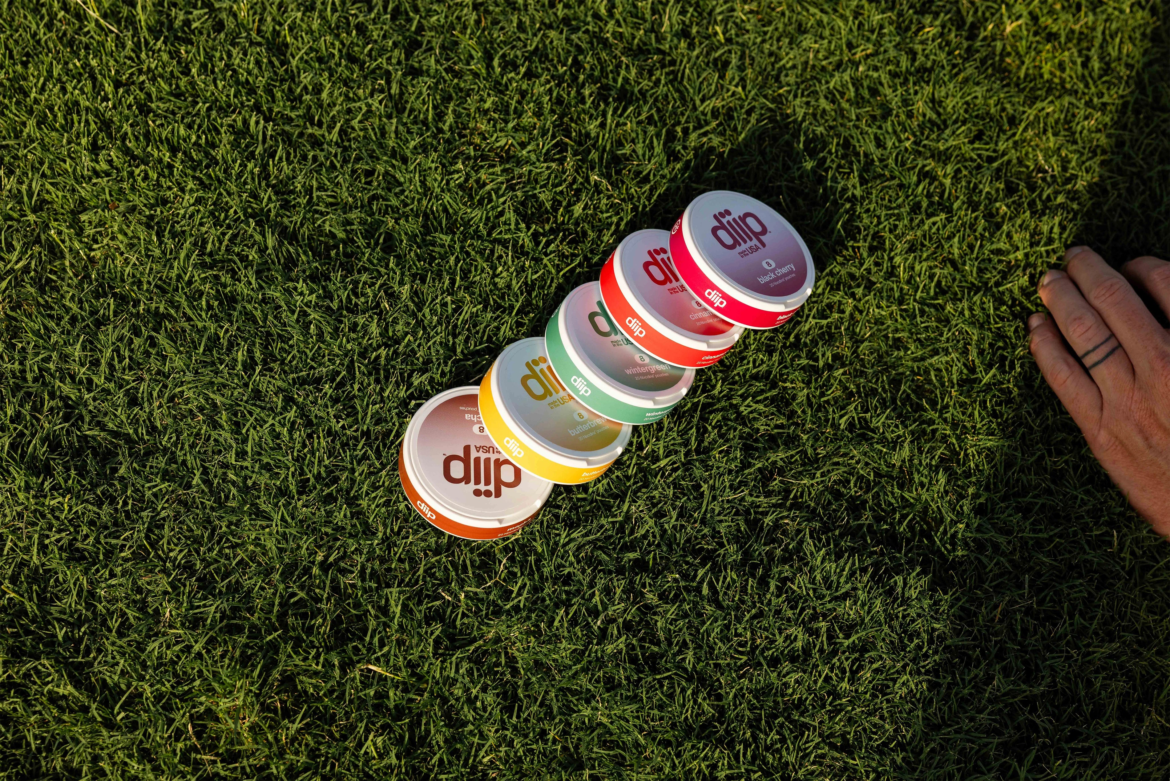 Collection of 'diip' branded nicotine-free dip pouch cans on grass with a hand placed next to them