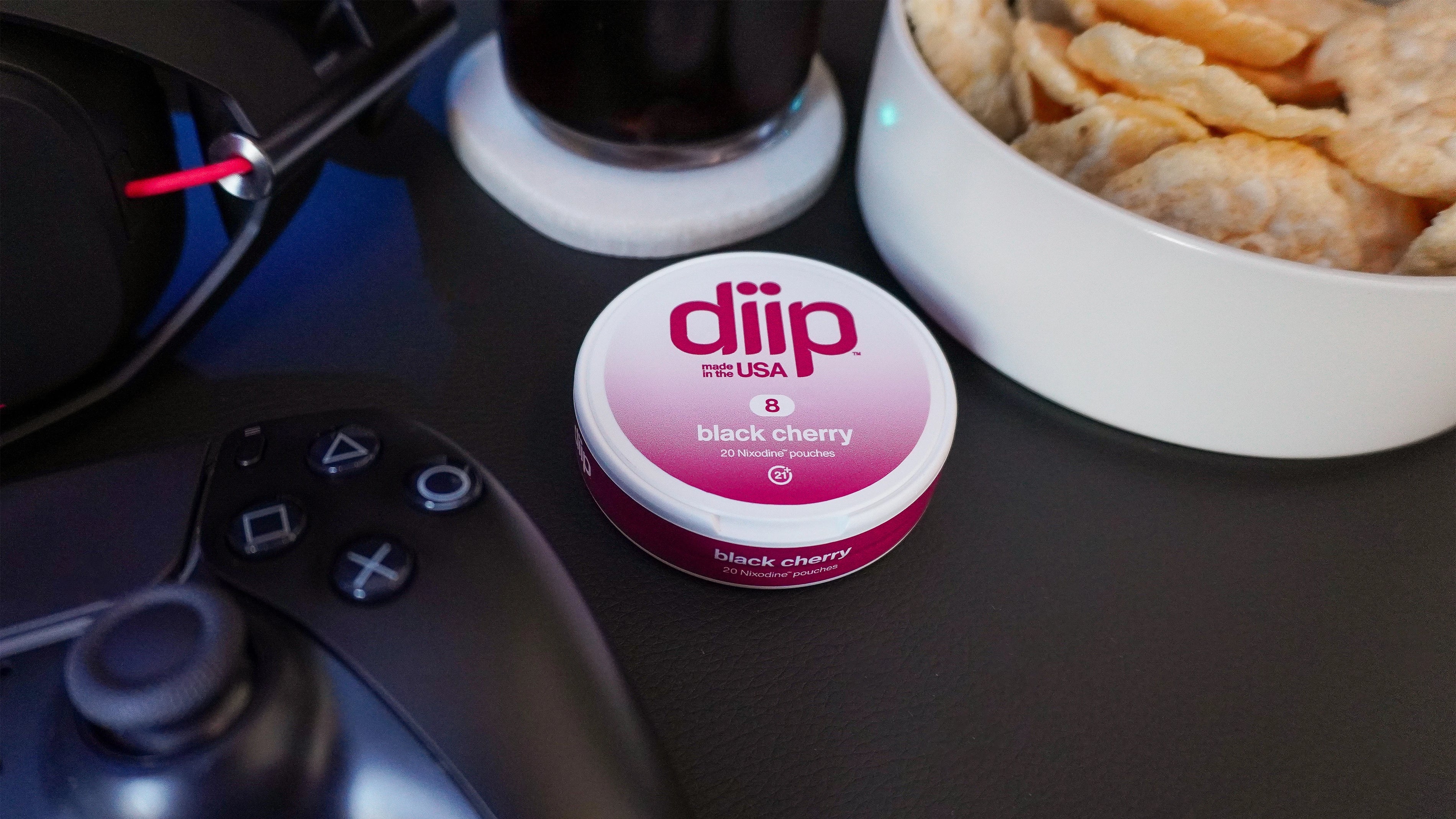 'diip' branded 8 black cherry nicotine-free dip pouch can laying on a gaming desk with a gaming controller, headphones, and snacks